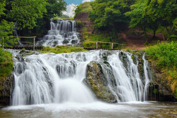 Fototapeta premium Dzhurynskyi waterfall cascades over tiered rocks into a foamy stream, surrounded by lush green trees and mossy stone in Ternopil region, Ukraine, on a bright summer day