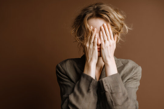 Stressed woman hiding face in hands against brown backdrop, conveying overwhelm, anxiety, or emotional distress in a relatable, raw portrait