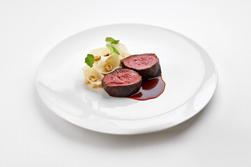 Filet Mignon sliced into two portions, cooked to a perfect medium-rare. The meat is elegantly garnished with white vegetables, hazelnuts, and microgreens, arranged artistically on a white plate.