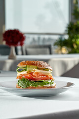 A classic bagel sandwich filled with smoked salmon, cream cheese, lettuce, cheese, and cucumber, served on a white plate against a bright white background with strong shadows.