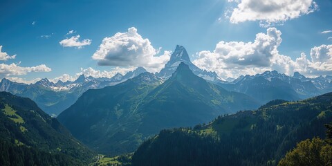 A panoramic view of the high Alps mountains with lush green forests beneath a cloudy blue sky, emphasizing seasonal change