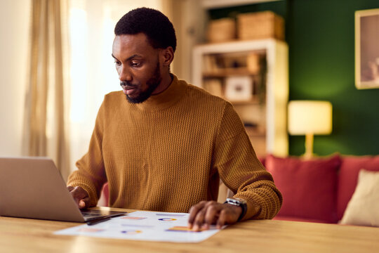 Focused Man Working From Home On Laptop With Documents And Charts On Wooden Desk