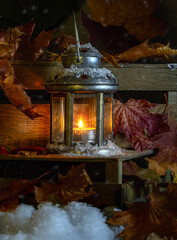 A burning candle in a small lantern against a background of fallen leaves and snow. In anticipation of Christmas and the New Year.