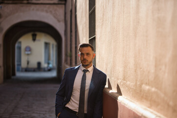 Young, handsome, dark-haired businessman wearing a suit and tie, leaning against a wall with a serious expression, smoking. The man is the director of a multinational company.