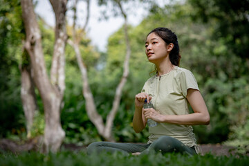 A young Asian woman opens a plastic water bottle on the grass in a calm park, while looking for her friend, having just finished a morning workout, surrounded by serene greenery, trees, and nature