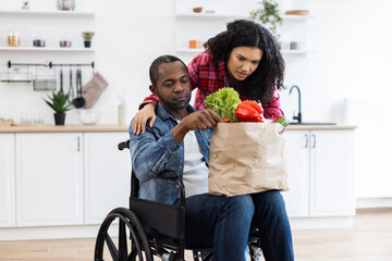 A Black couple in a modern kitchen, one in a wheelchair, unpacking groceries together, promoting inclusivity and healthy eating.