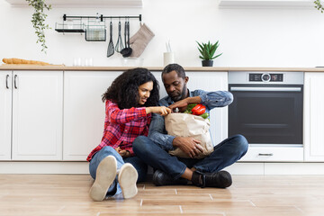 A couple sits on the floor in their kitchen, unpacking a paper grocery bag filled with fresh produce.
