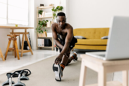 Young african man doing a stretching exercise on the floor, following an online workout program on a laptop in his living room