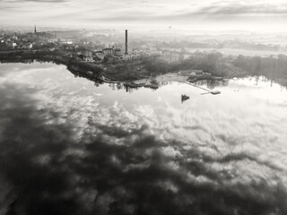 Czarno-białe zdjęcie lotnicze Opola przedstawiające zalane wyrobisko Kamionka Bolko o świcie, z widocznym w oddali kominem dawnej cementowni, panoramą miasta oraz efektownymi chmurami odbijającymi się