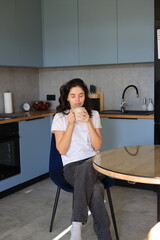 Woman sitting in the kitchen with closed eyes, holding a cup of coffee with both hands and enjoying a calm, peaceful moment at home