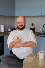 Man showing a negative facial expression while holding a bitten apple, reacting to an unpleasant or sour taste in a home kitchen setting