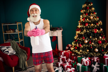 Fototapeta premium Elderly man dressed as Santa Claus cleaning and preparing near a decorated Christmas tree at home for the holidays