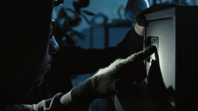 Two male thieves trying to guess multi-digit code cracking electronic safe, man with headlamp pressing buttons while accomplice in balaclava aiming flashlight at them