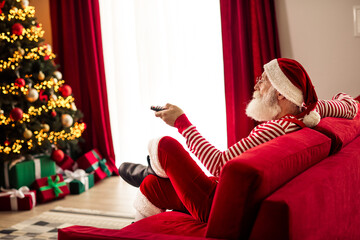 Cheerful man dressed as Santa Claus relaxing at home with a television remote during Christmas...