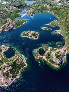 Aerial view of a vast, deep blue mountain lake system with numerous islands, rocky shores, and green vegetation under a bright, clear sky.