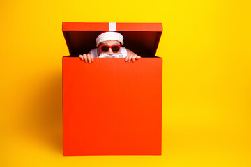 Santa in a red box smiling with sunglass in a bright yellow studio for a playful holiday gift scene