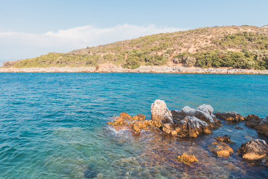 Rocky shoreline with clear blue water and hills under sunny sky