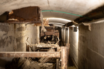 Deep perspective inside an abandoned cement factory tunnel showing the heavily rusted, collapsing remains of a conveyor belt system and machinery.