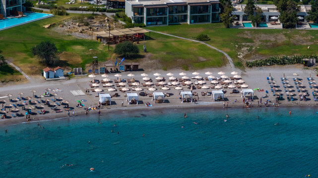 Aerial view of sun-kissed beach with vibrant parasols contrasting against the deep blue sea and modern architecture, Kolympia, Rodos, Greece.
