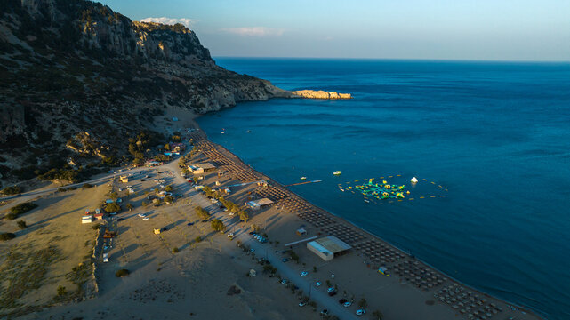 Aerial view of a sun-kissed beach meeting the turquoise sea, shadowed by rugged cliffs and dotted with sunbeds, umbrellas and floating platforms, Kolympia, Rodos, Greece.