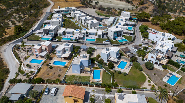 Aerial view of modern white villas with azure pools nestled amidst the sun-drenched landscape, a stark contrast to the arid hills, Kolympia, Rodos, Greece.