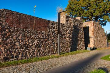 Strzelce Krajeńskie (niem. Friedeberg in der Neumark) – oświetlony słońcem kamienny mur na Starym Mieście i brukowana ścieżka z nowoczesną latarnią uliczną w otoczeniu parku.