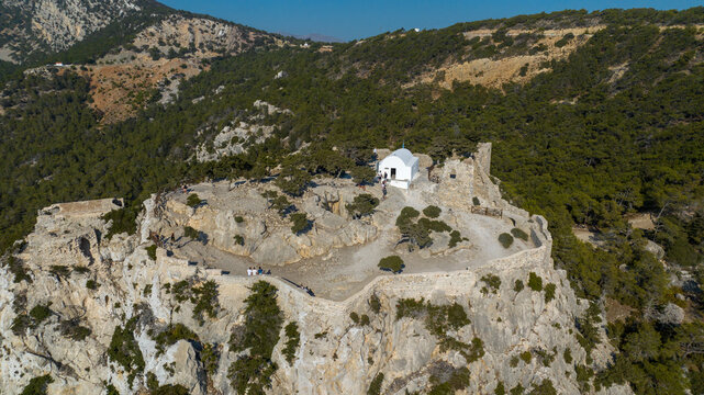 Aerial view of a white chapel perched atop a rugged, rocky cliff, embraced by verdant trees, creating a striking contrast against the azure sky, Rhodes, Greece.