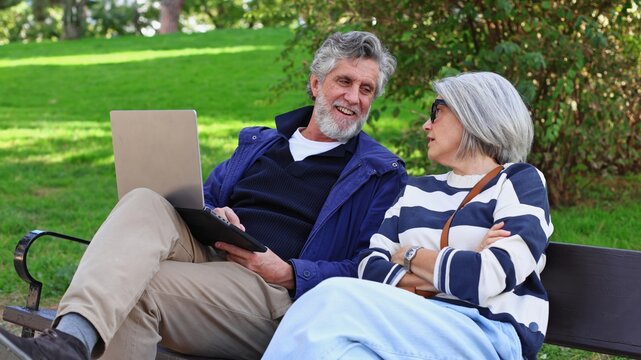 Senior couple relaxing on park bench using laptop