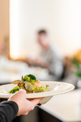 Naklejka premium A waiter's hands serve an elegantly plated gourmet dish of seafood, broccoli, and mushrooms in a fine dining restaurant setting with a shallow depth of field