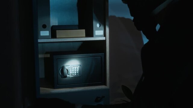 Male burglar with flashlight opening wooden cabinet, cracking metal safe box after second try, stealing cash from it and noticing gold bar