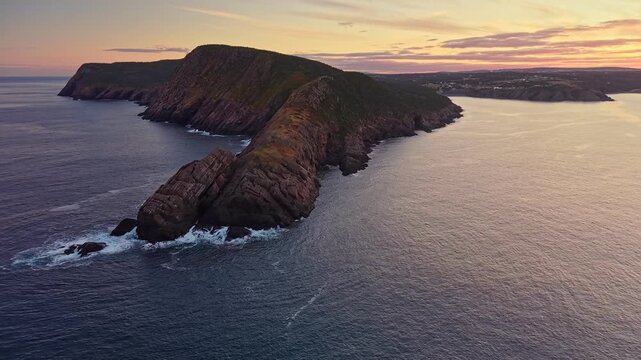 Wide aerial view reveals Torbay Point peninsula between two inlets, with rugged slopes, sheer cliffs and pastel sky stretching to the horizon.