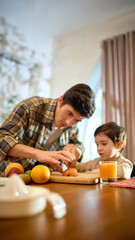 Orange juice glass in bright kitchen scene with lively warm father and little boy moment. Concept of lifestyle storytelling, health visuals, food promotion and positive emotional branding.
