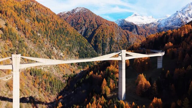 Aerial view of the Ganter Bridge piercing through the vibrant autumnal forest, contrasted against the snowy mountain peaks, Ried-Brig, Wallis, Switzerland.