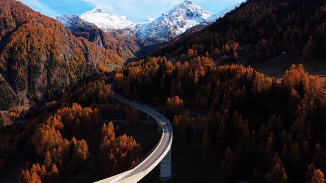 Aerial view of the Ganter Bridge winding through vibrant autumn trees and towards snow-capped mountains, creating a stark contrast, Ried-Brig, Wallis, Switzerland.