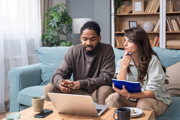 Young couple working from home in a cozy living room. He explains ideas at a laptop while she takes notes with a notebook. Genuine remote collaboration, coffee, casual lifestyle. Freelance work