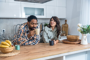Cheerful couple enjoying a relaxed coffee chat in the kitchen, leaning on the counter, smiling and talking about plans, cozy home morning vibe