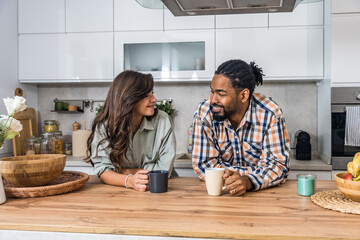 Cheerful couple enjoying a relaxed coffee chat in the kitchen, leaning on the counter, smiling and talking about plans, cozy home morning vibe