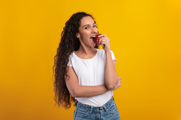 A smiling young woman stands against a yellow wall, holding a red apple. She is happily taking a bite, promoting healthy eating and nutrition. Her cheerful demeanor shines through.