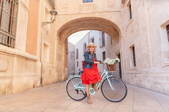 Senior woman wearing red dress and straw hat posing with bicycle in Valencia city center - Powered by Adobe