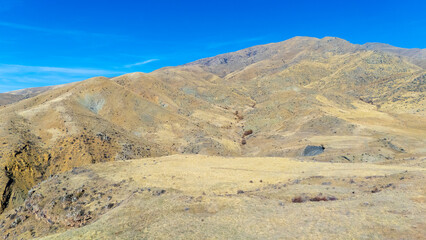 Expansive aerial view of dry rolling mountains under a clear blue sky. Layered ridges stretch into the distance with soft haze, creating a dramatic and natural panoramic land