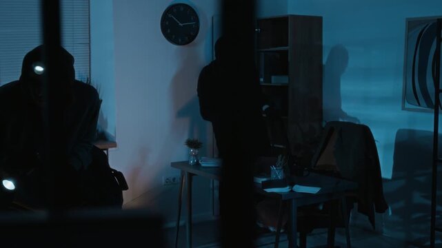 Young male burglar taking jacket from leather chair and putting it on when his accomplice examining office using flashlight during robbery