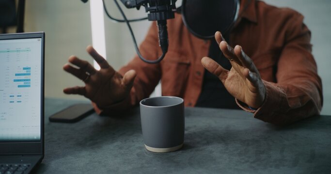 Close Up of Expressive Male Hands Gesturing While Discussing Ideas in Podcast Studio. Hands Visible as Man Talks, With Coffee, Laptop, Smartphone on Table, Laptop Screen Showing Analytics Dashboard.