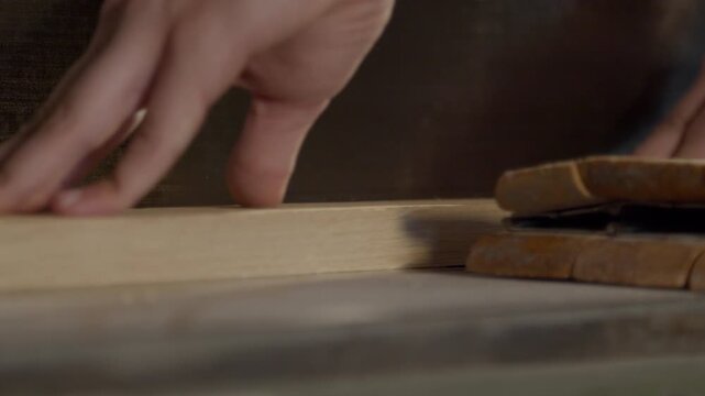 High quality video of a hand guiding a wooden board across a saw in warm cinematic light. Slow motion captures dust, grain and precise woodworking in a calm workshop atmosphere.