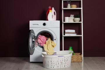 Modern washing machine, detergents and clothes near burgundy wall indoors
