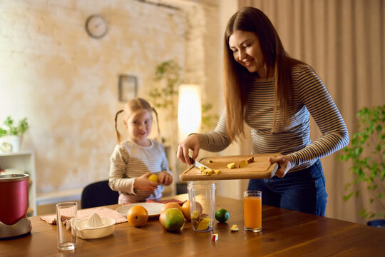 Mother and daughter preparing smoothie ingredients with orange juice beside blending container in warm kitchen. Concept of beverage creation, healthy nutrition, lifestyle and cheerful emotion.
