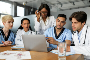 Fototapeta premium Concentrated multiethnic team of doctors men and women in workwear sitting at table at clinic, having breefing, looking for solutions, discussing clinical cases, looking at laptop screen and chatting