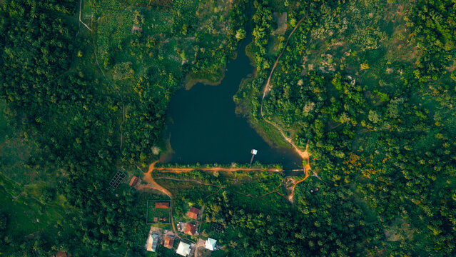 Aerial view of a dark lake embraced by a vivid green forest with a small bridge and building by the water's edge, Erin Ile, Kwara, Nigeria.
