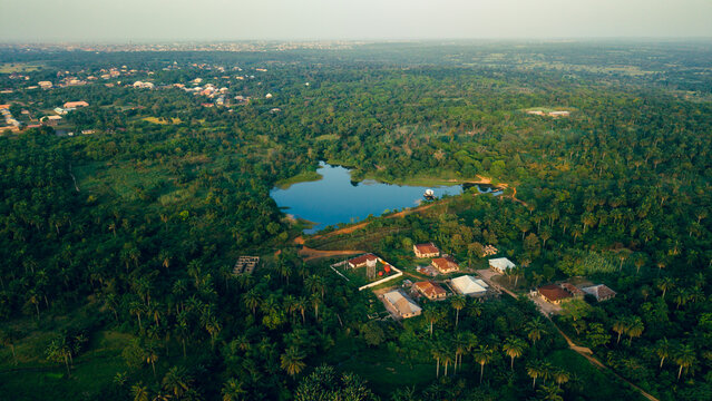 Aerial view of a serene lake nestled amidst a vibrant green forest, with buildings peeking through the trees, Erin Ile, Kwara, Nigeria.