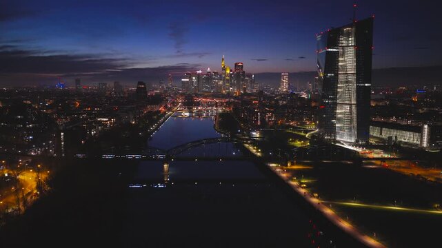 Aerial view of the ECB and Frankfurt skyline at dusk, a vibrant mix of illuminated skyscrapers and the dark river Main, Frankfurt am Main, Hessen, Germany.
