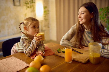 Little girl drinking fresh smoothie while mother watches with warm joyful emotion. Concept of healthy drinks, cozy lifestyle visuals and emotional family branding.